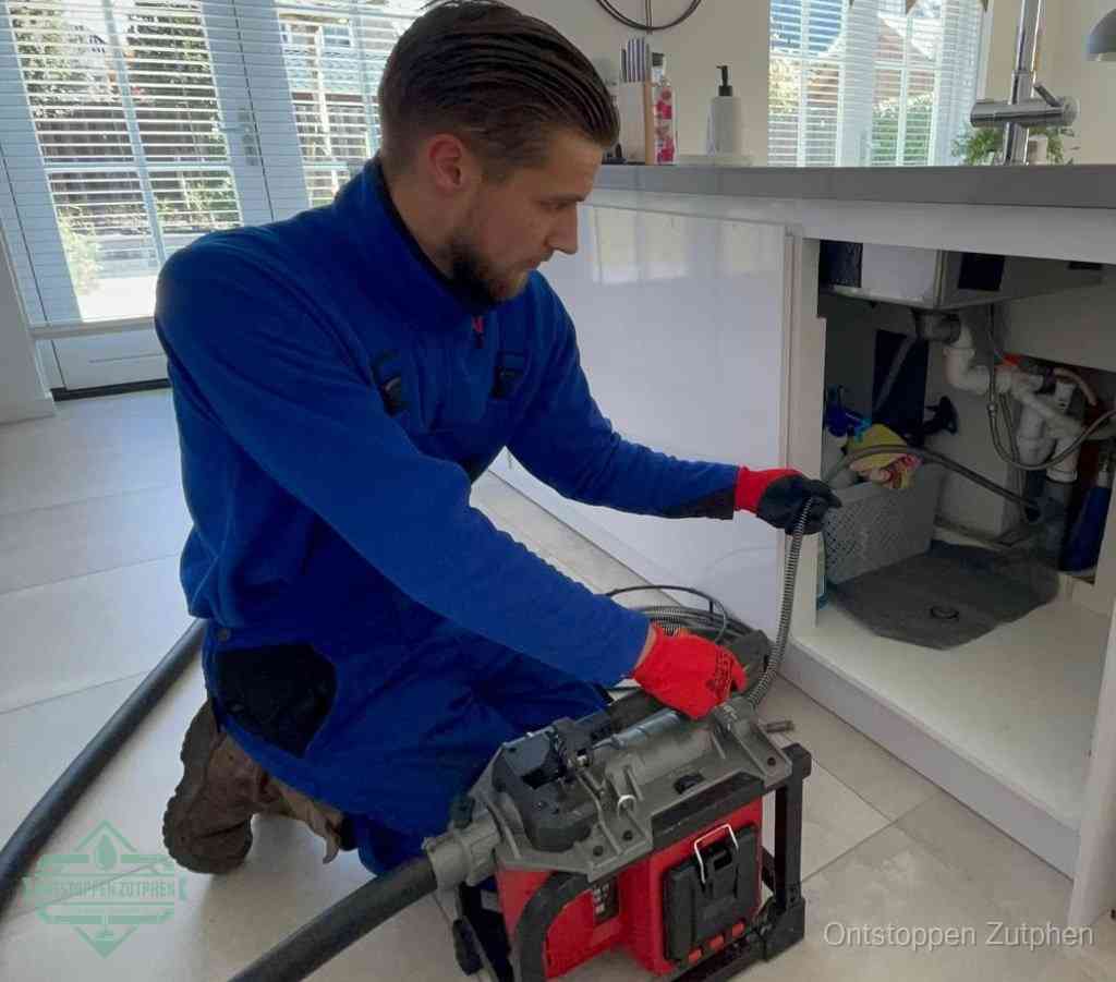 Een monteur met rode handschoenen knielt in een keuken en voert een elektrische ontstoppingsveer in tijdens het professioneel afvoer ontstoppen in Zutphen